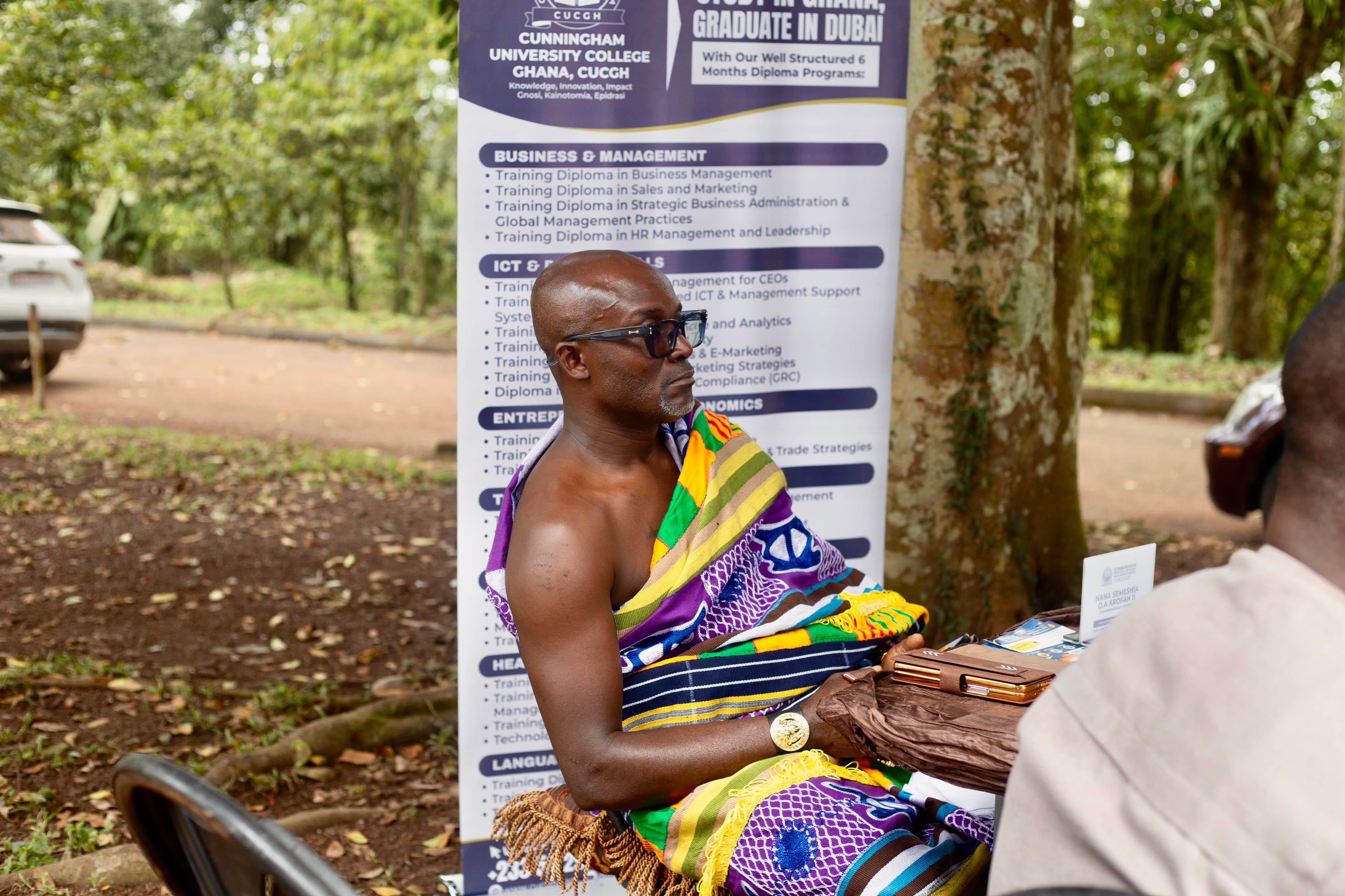 Nana Semeshia Ohene Asah Krofah II in colourful traditional cloth seated at the meeting table beside a programme banner.